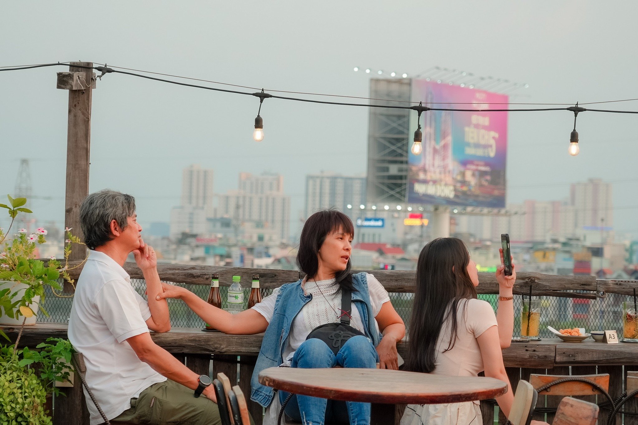 Three people are seated at an outdoor rooftop venue in Thao Dien, Saigon. The scene features a wooden railing, string lights with exposed bulbs, and a