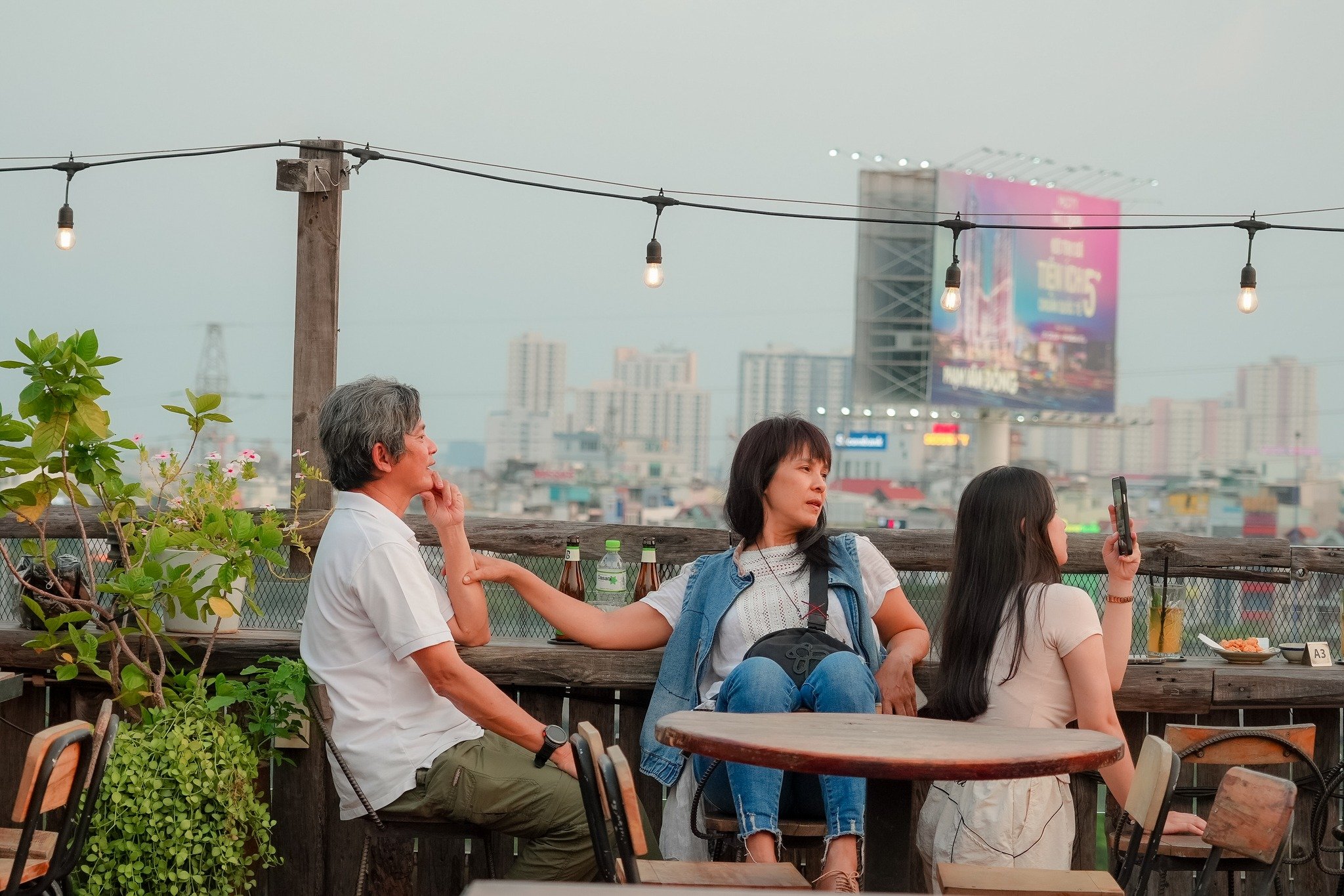 Three people are seated at an outdoor rooftop venue in Thao Dien, Saigon. The scene features wooden railings, string lights, potted plants, and a city
