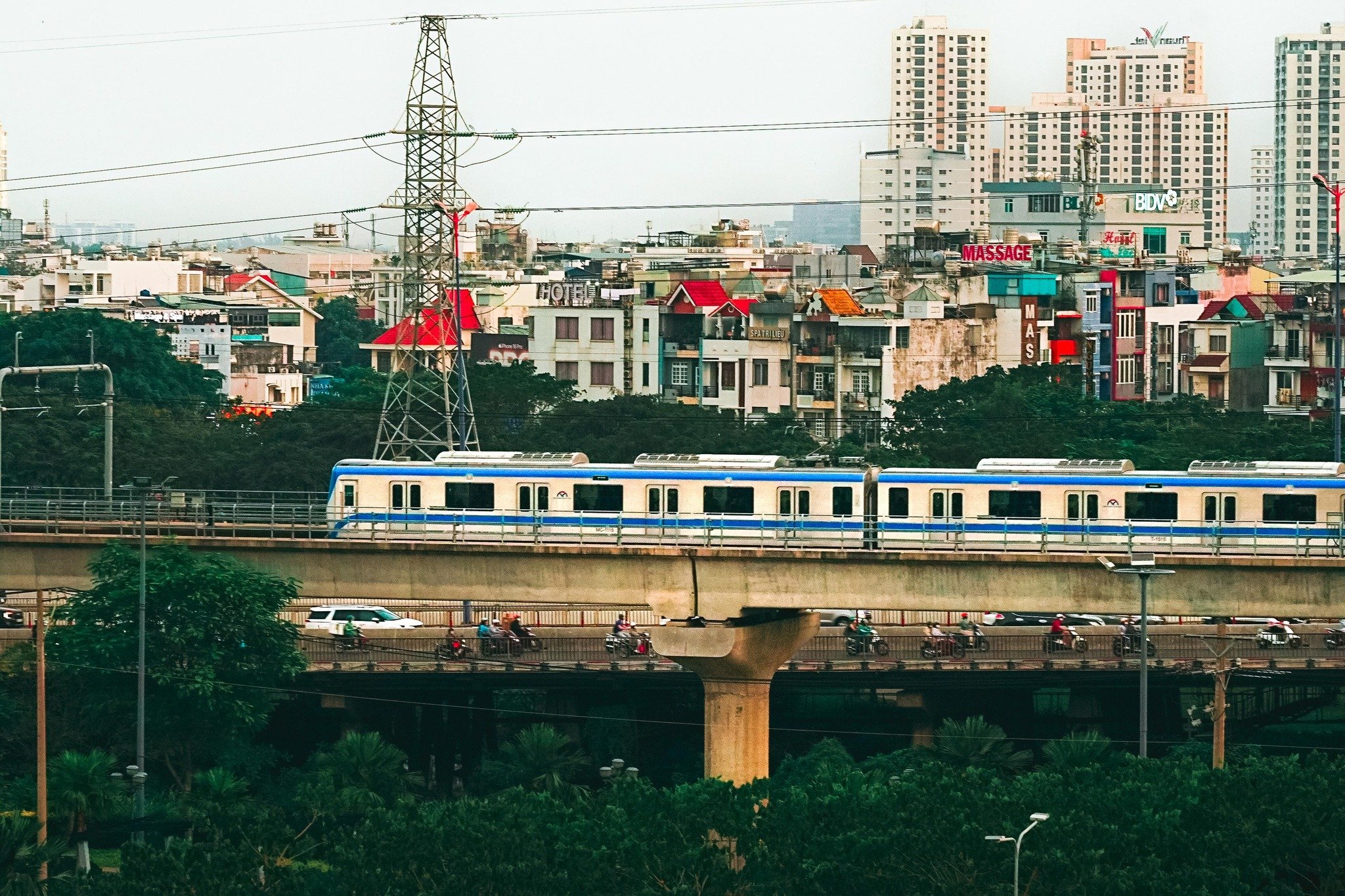 A vibrant urban scene in Thao Dien, Saigon, featuring a blue and white elevated train on a concrete viaduct. Below the train, a busy road with motorbi