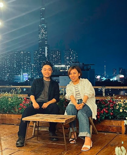 Two people seated on a rooftop terrace with a wooden table, surrounded by flower planters. Background features a nighttime cityscape with illuminated 