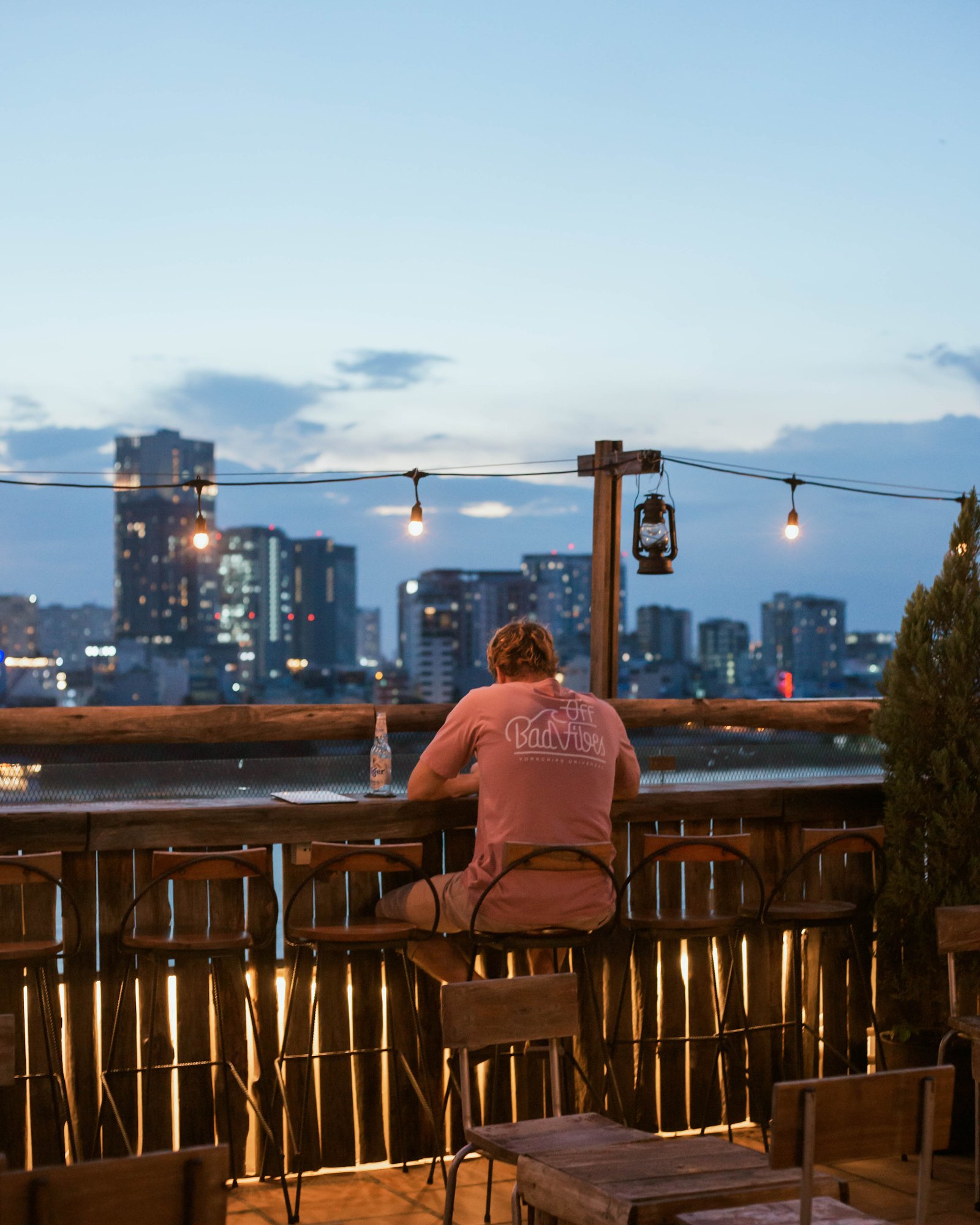 A person in a pink 'Off Bad Vibes' shirt sits on a wooden bar stool at a rooftop venue overlooking Saigon's cityscape during twilight. The scene inclu