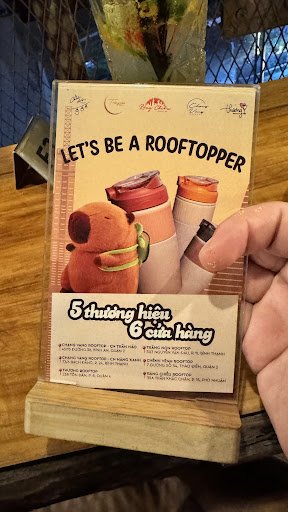 A hand holding a promotional menu card titled 'LET'S BE A ROOFTOPPER' with illustrations of a bear and beverage containers. The card features text in 