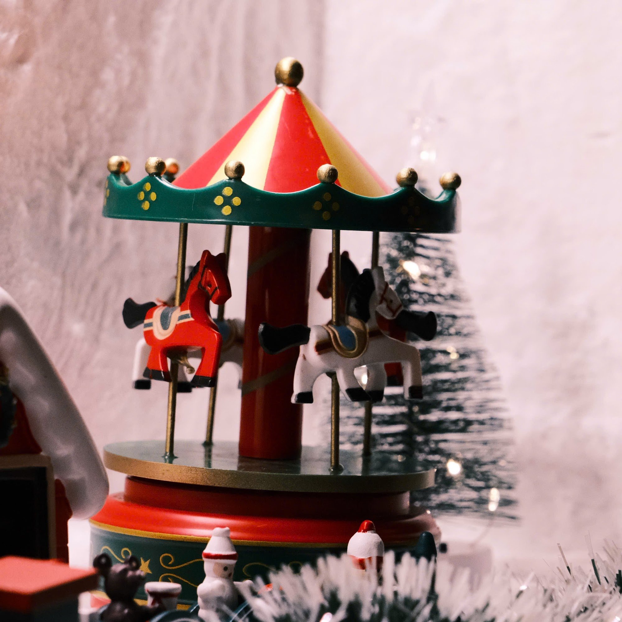 A close-up of a miniature carousel with red and white horses under a red and yellow striped canopy, featuring green base with gold accents. The backgr