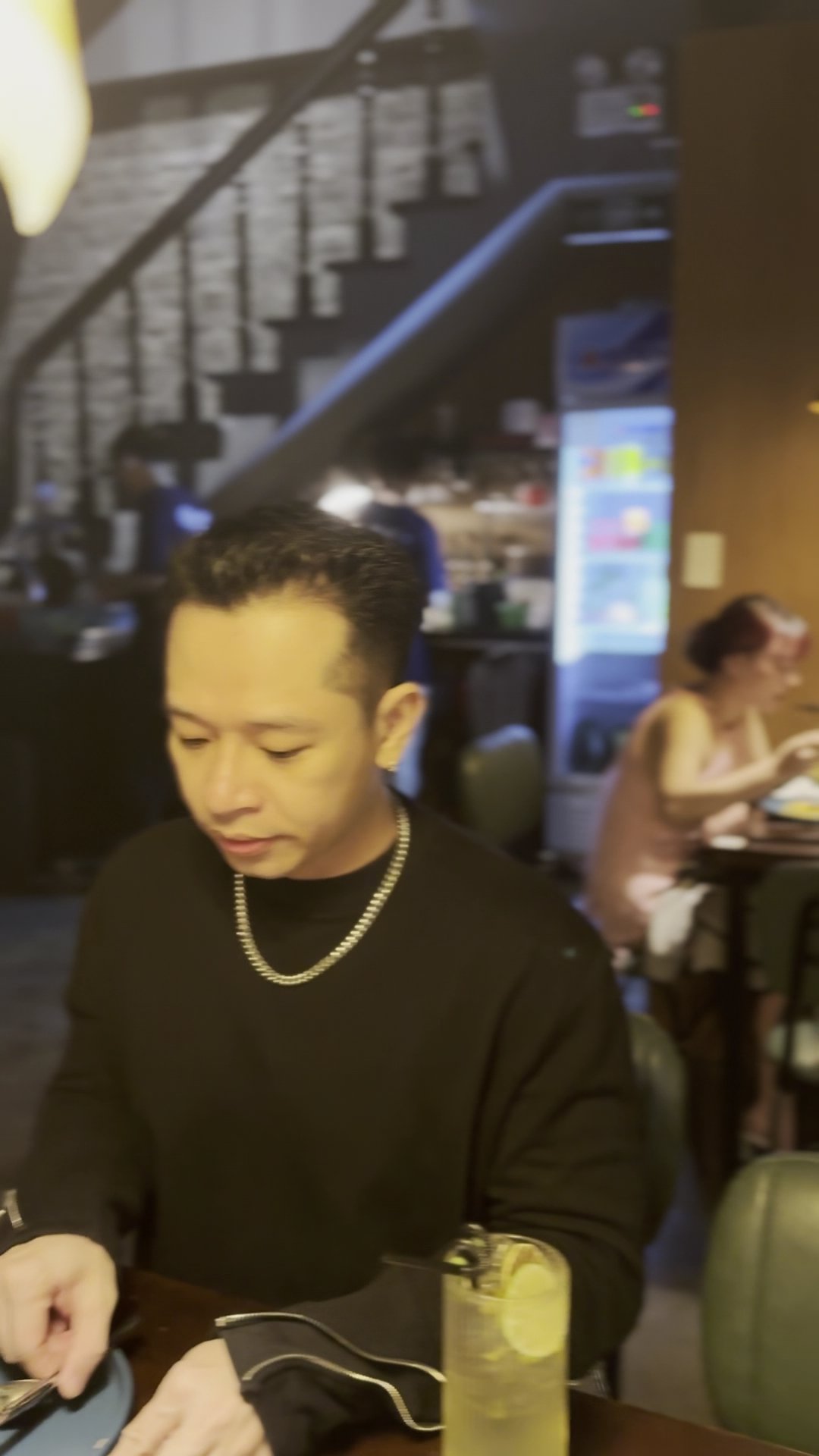A man in a black sweater and gold chain sits at a table in a restaurant. On the table, there's a glass with a lemon slice and a drink. Background feat