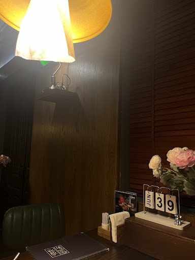 A dimly lit interior space with wooden paneling on the walls. Features include a desk with a dark folder, a green chair, a wall-mounted lamp with a ye