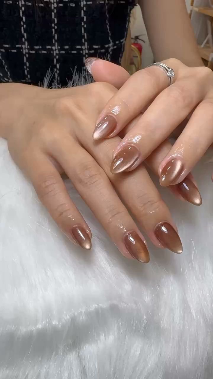 A close-up of a person's hands displaying well-manicured nails painted in a glossy metallic rose gold color with some glitter on the tips, resting on 