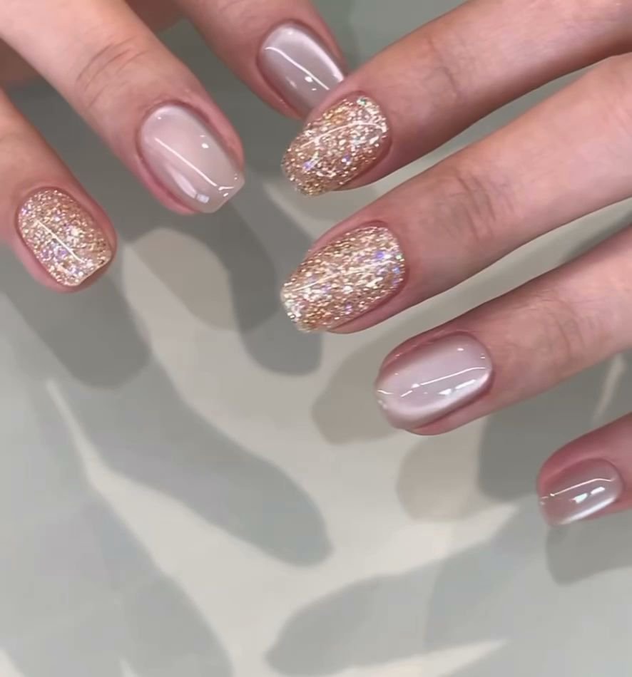 A close-up of a person's nails adorned with glittery gold and nude polish against a blurred background that suggests an indoor setting, possibly in Th