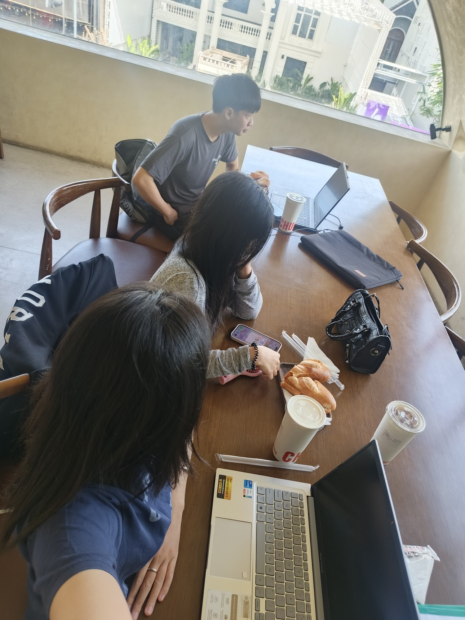 A group of three people (two women and one man) are gathered around a wooden table in a café or co-working space. On the table, there are two laptops,