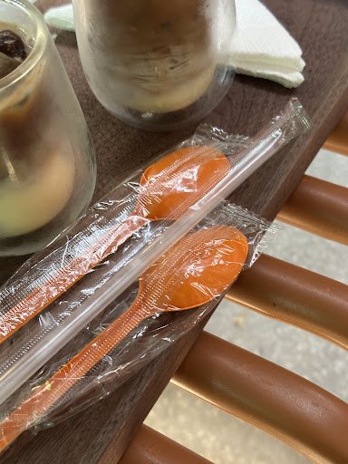 The image shows a close-up of two orange plastic spoons in clear packaging on top of a wooden surface with what appears to be an iced coffee drink par