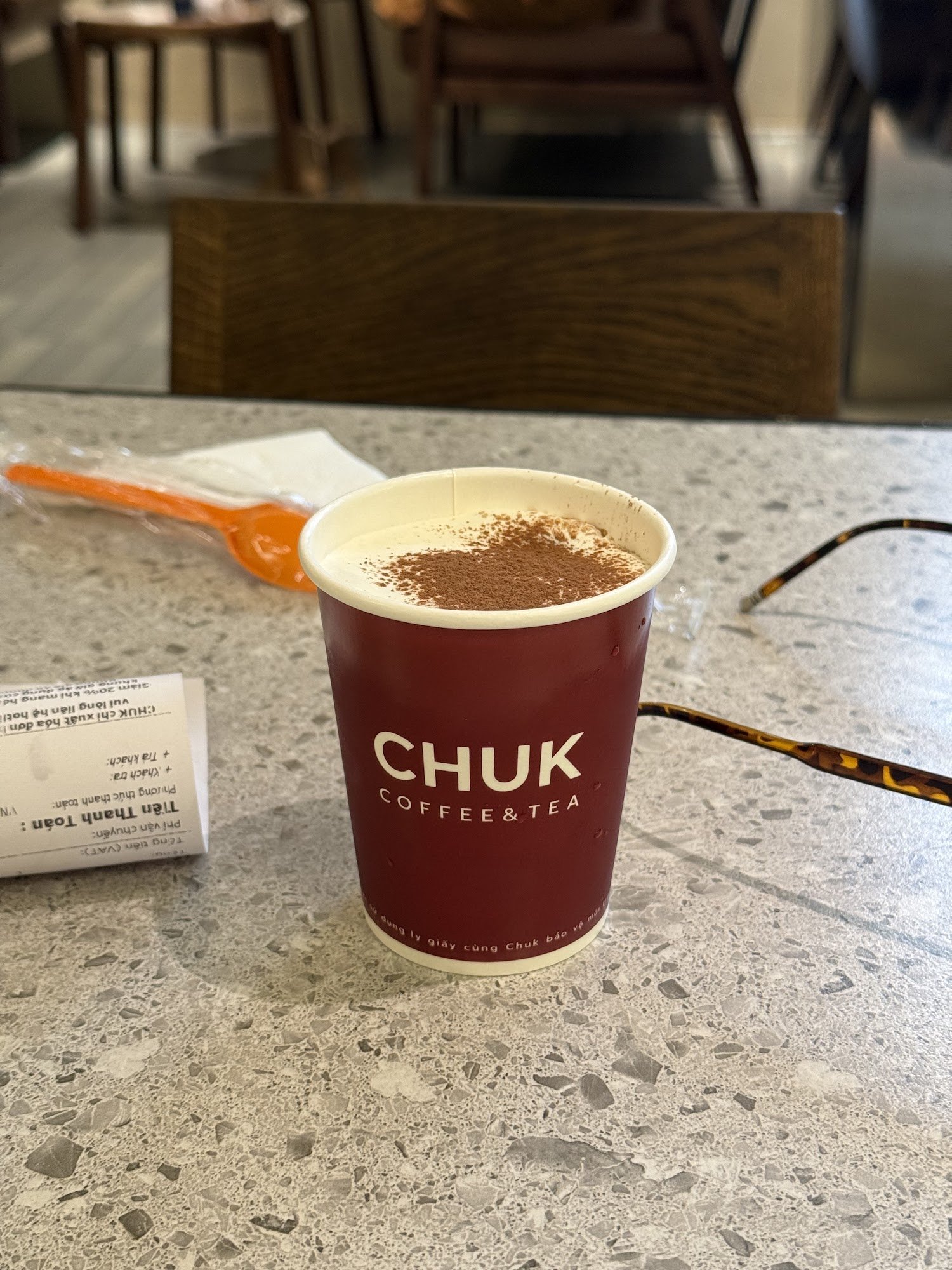 A cup of coffee from CHUK Coffee & Tea on a marble countertop in what appears to be an indoor setting with wooden chairs and tables visible in the bac