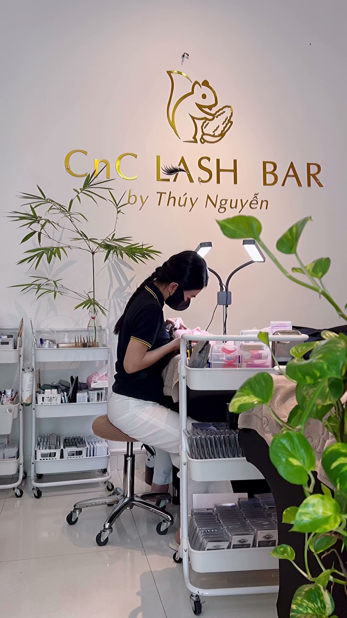 The image shows an interior view of a professional lash bar named 'CNC LASH BAR by Thúy Nguyêñ'. A technician is seated at her workstation, surrounded
