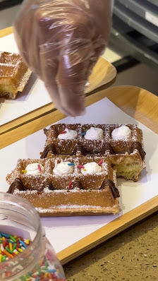 A close-up of a dessert consisting of waffles topped with chocolate sauce and powdered sugar, garnished with whipped cream dollops and colorful sprink