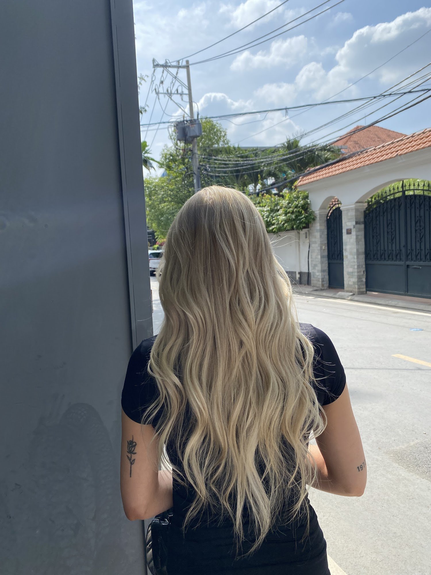 A person is standing on a street corner in an urban area during the daytime. The individual has long blonde hair and tattoos visible on their arms, we