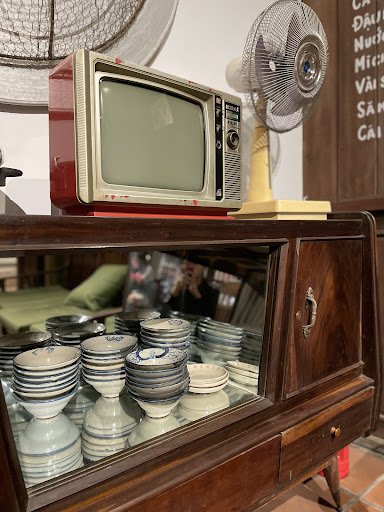The image shows a vintage-style wooden cabinet displaying stacks of ceramic dishes and bowls on its shelves. On top of the cabinet, there is an old-fa