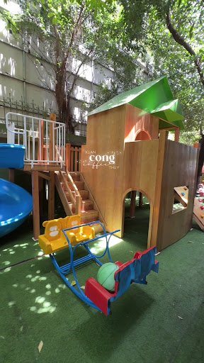 The image depicts an outdoor playground in Thao Dien, Saigon. It features a wooden playhouse with a green roof, a blue slide, a yellow and blue swing 