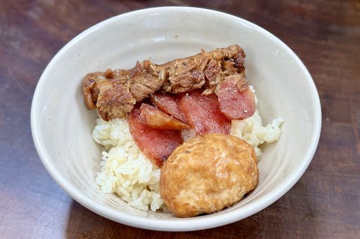 A bowl containing a meal that includes rice topped with slices of salami and pieces of meat, alongside what appears to be a fried biscuit or dumpling.