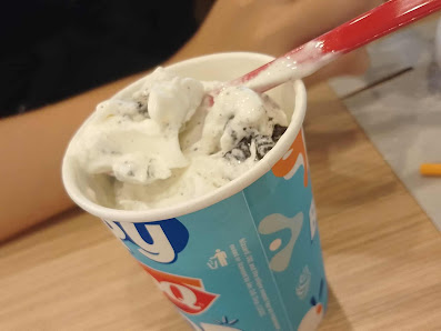 A close-up of a cup containing ice cream or frozen dessert topped with what appears to be chocolate chips and possibly nuts, held by someone using a r