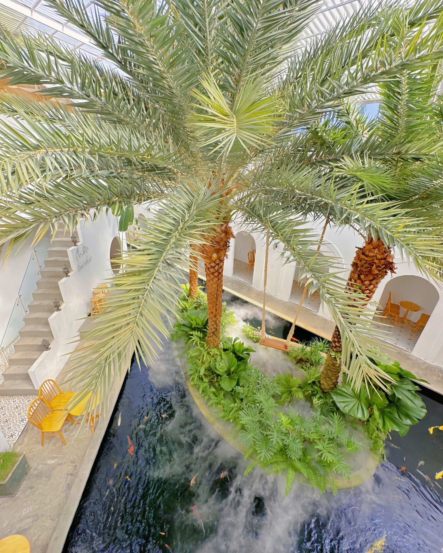The image depicts an outdoor venue in Thao Dien, Saigon, featuring a central water feature with koi fish, surrounded by lush tropical plants including