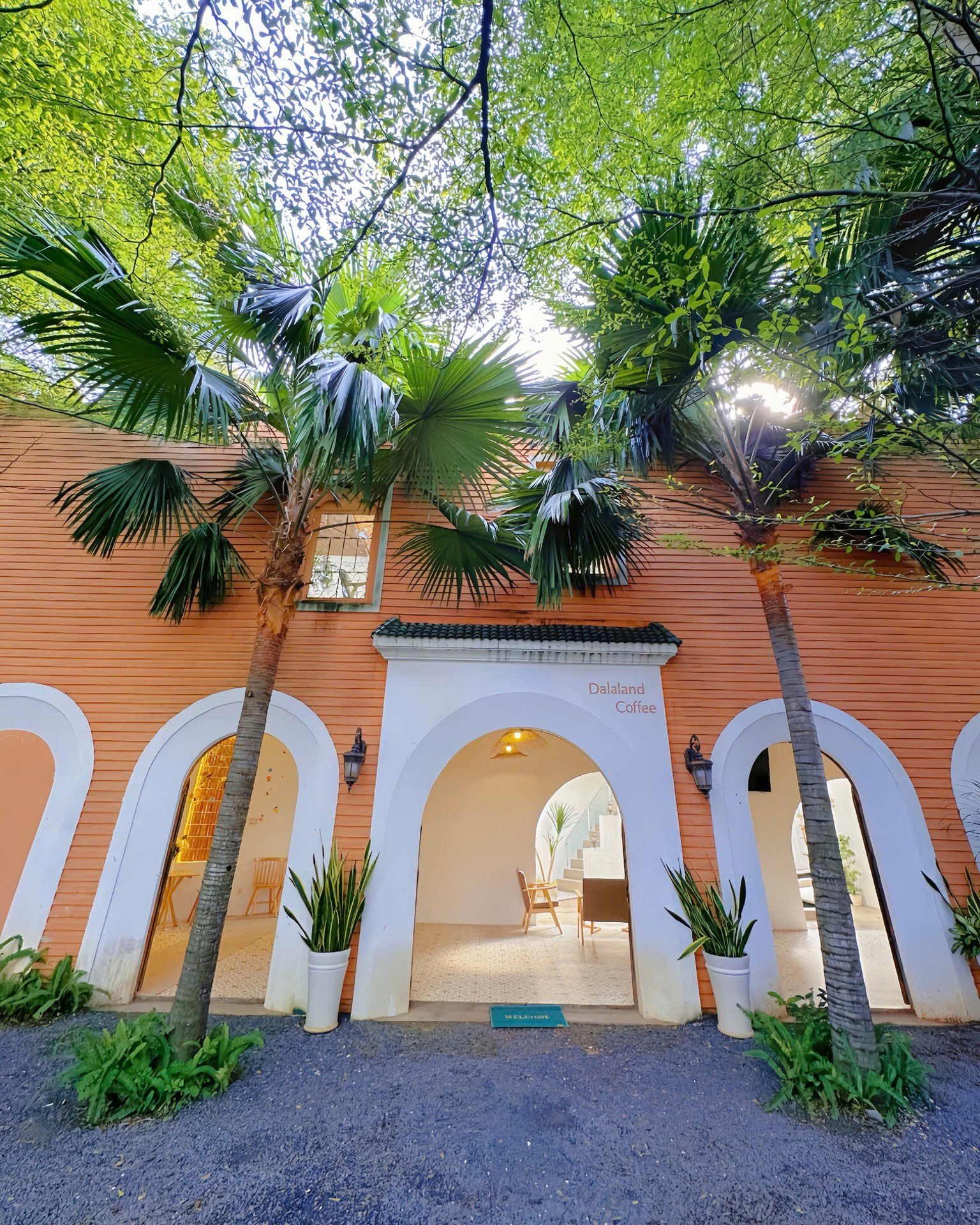 The image depicts the exterior of Dalaland Coffee, a venue in Thao Dien, Saigon. The building features a warm orange facade with white arched doorways
