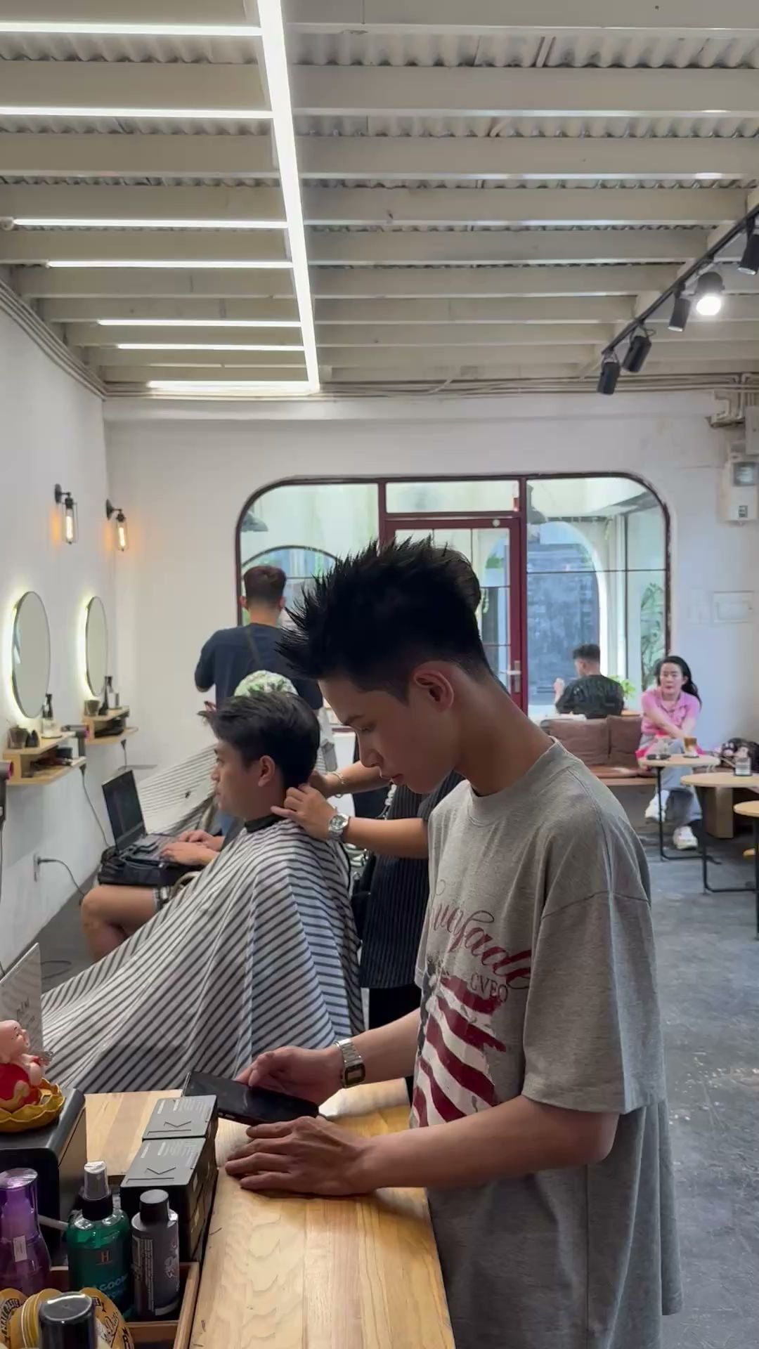 The image shows an interior view of a modern barber shop or hair salon where customers are receiving services and other patrons are waiting in the bac