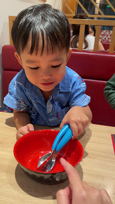 A young child is seated at a red booth in an indoor dining area, interacting with someone off-camera who is holding out a plate and utensils.
