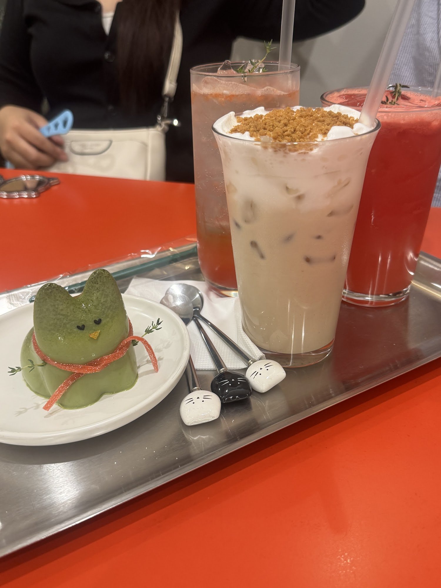 A green dessert shaped like a cat with red decorative elements around it. Accompanied by two beverages in glasses and another drink in a cup on the tray.