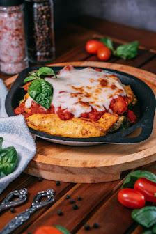 A baked dish topped with melted cheese and basil leaves is presented on a black cast iron skillet placed on a wooden serving board, surrounded by fres