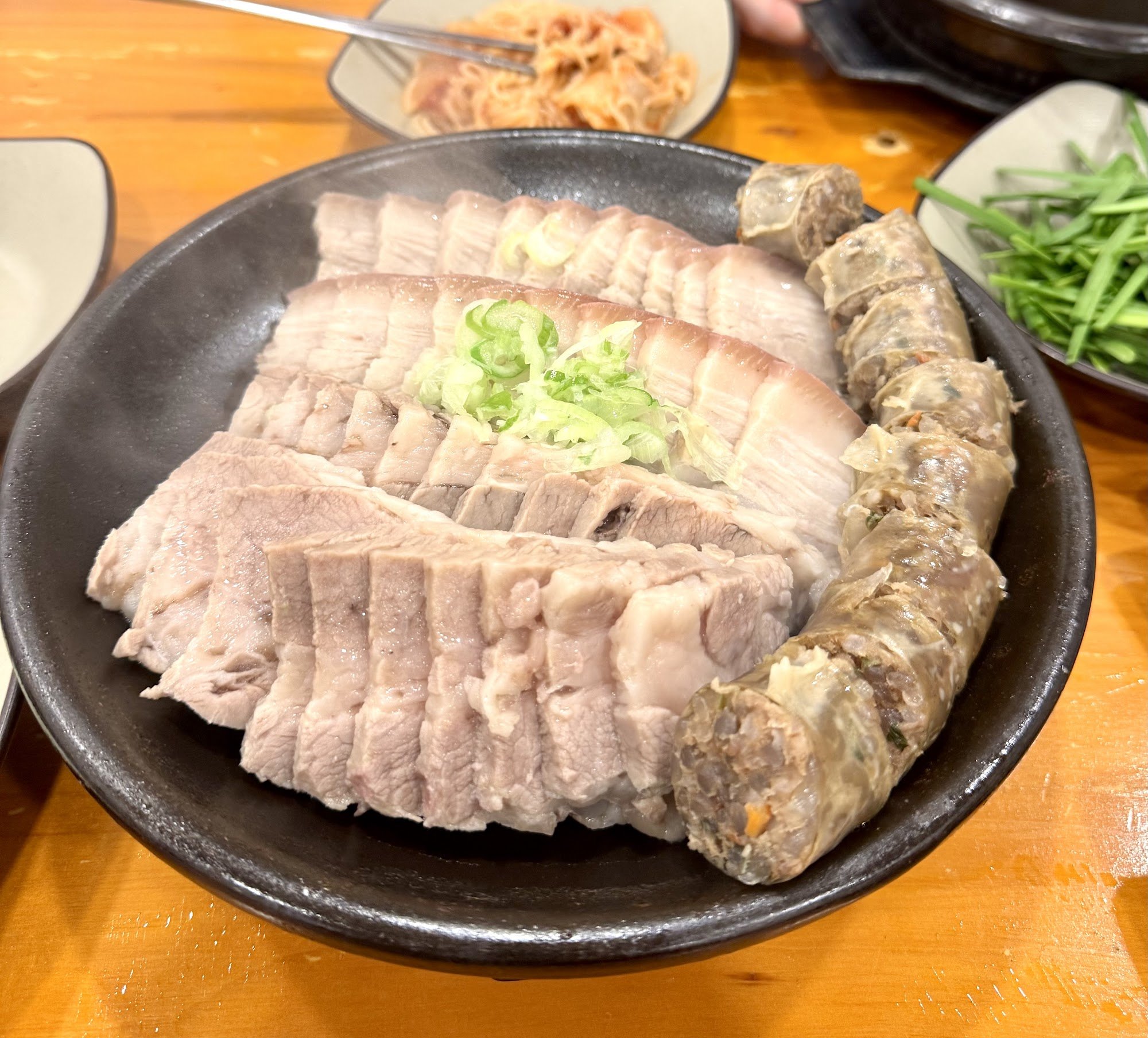 A close-up of a dish consisting of sliced pork belly and rolled meat on a black plate with garnishes such as green onions, likely served hot.