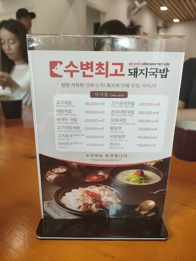 A menu stand displaying various food items and their prices in Vietnamese dong at a restaurant named 'COSAM'. The background shows part of the dining 