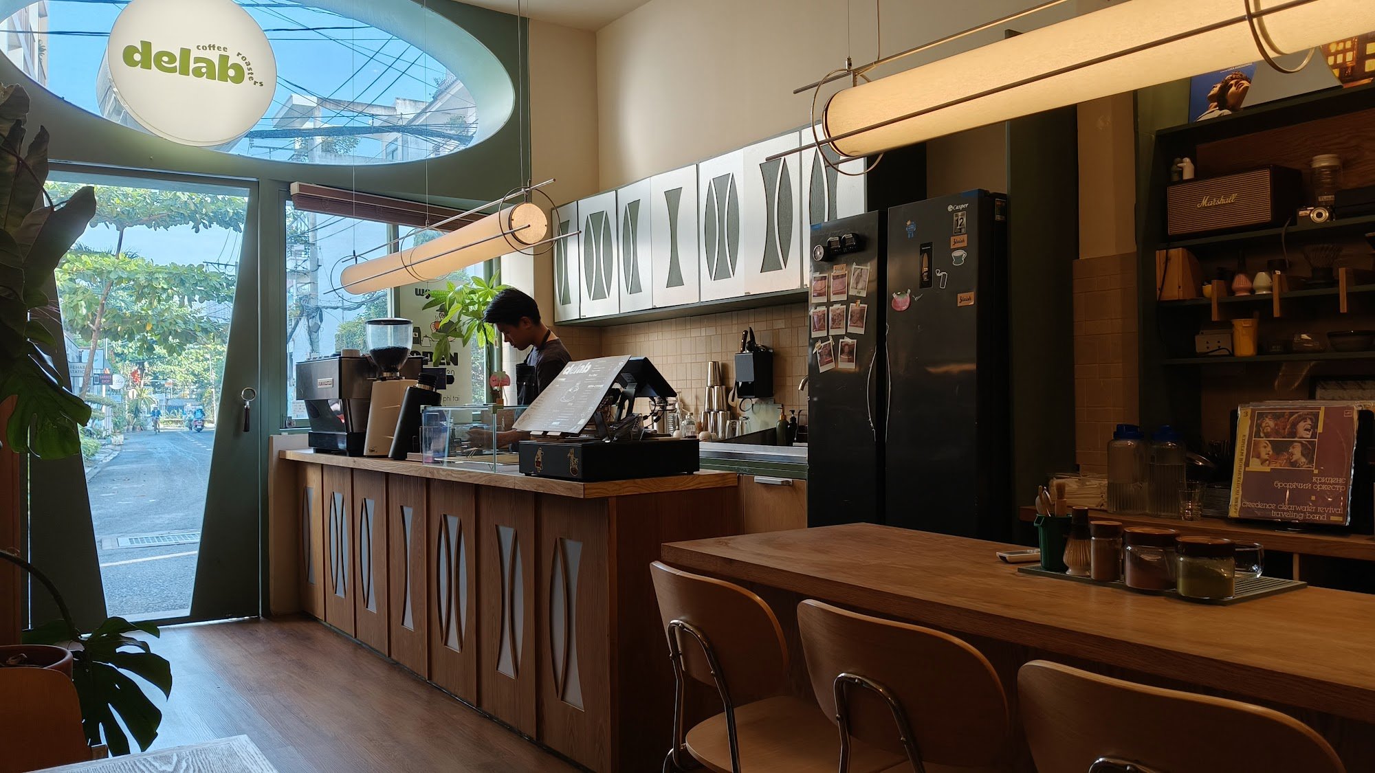 The image shows an interior view of a coffee shop named 'delab', which has wooden furniture and modern decor elements such as large windows, pendant l