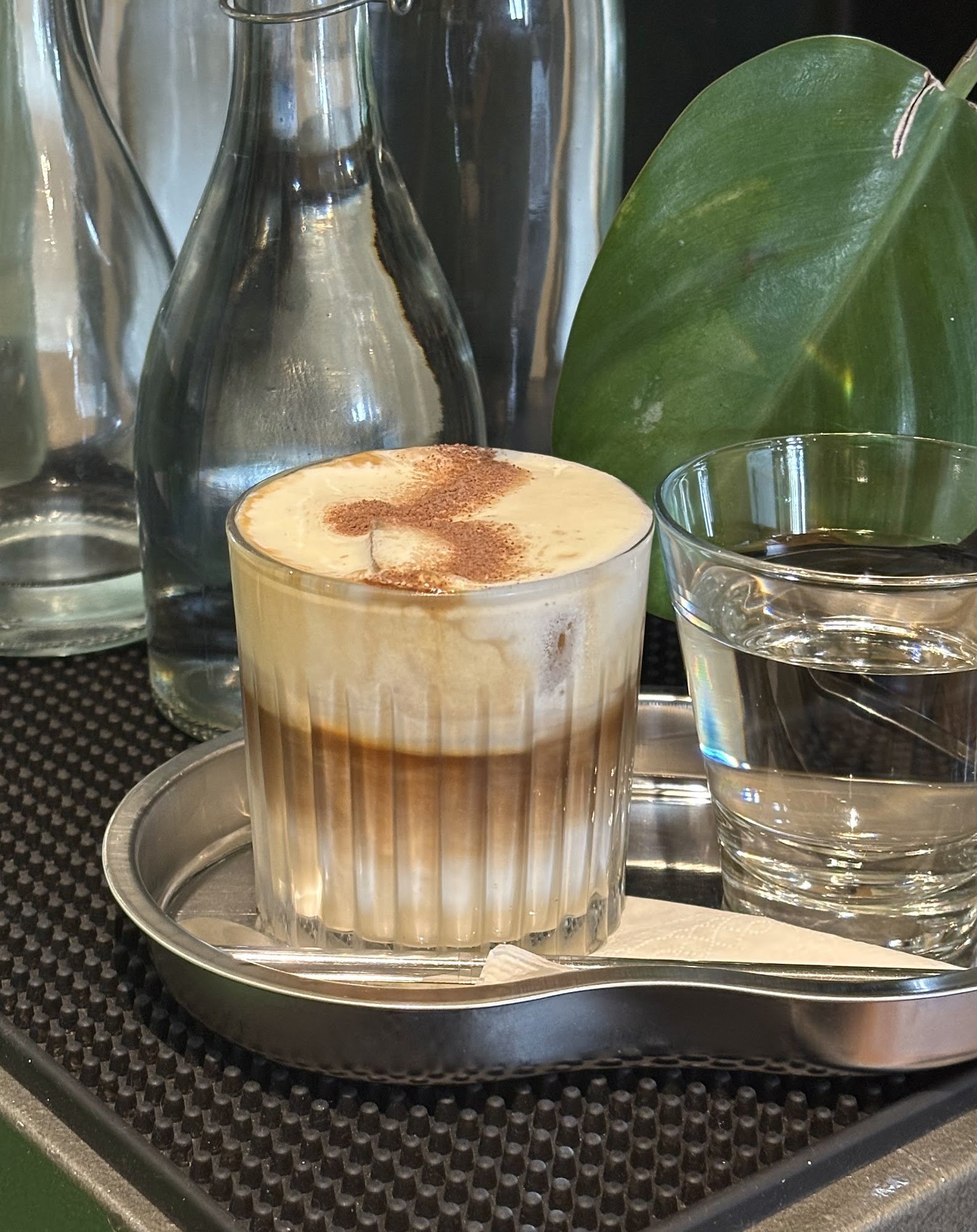 A layered coffee beverage in a glass on a metal tray, accompanied by a clear drinking glass with water and various bottles of liquids likely used for 