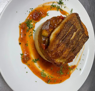 A plated dish featuring a piece of grilled bread topped with what appears to be caramelized onions or mushrooms in sauce, garnished with herbs and ser