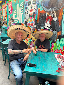 Two individuals wearing straw hats with 'BIER' text seated at turquoise outdoor tables. The venue features vibrant murals on the walls, including a la