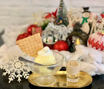 A festive dessert display featuring a scoop of vanilla ice cream in a glass bowl topped with whipped cream and a waffle cone, accompanied by a shot of