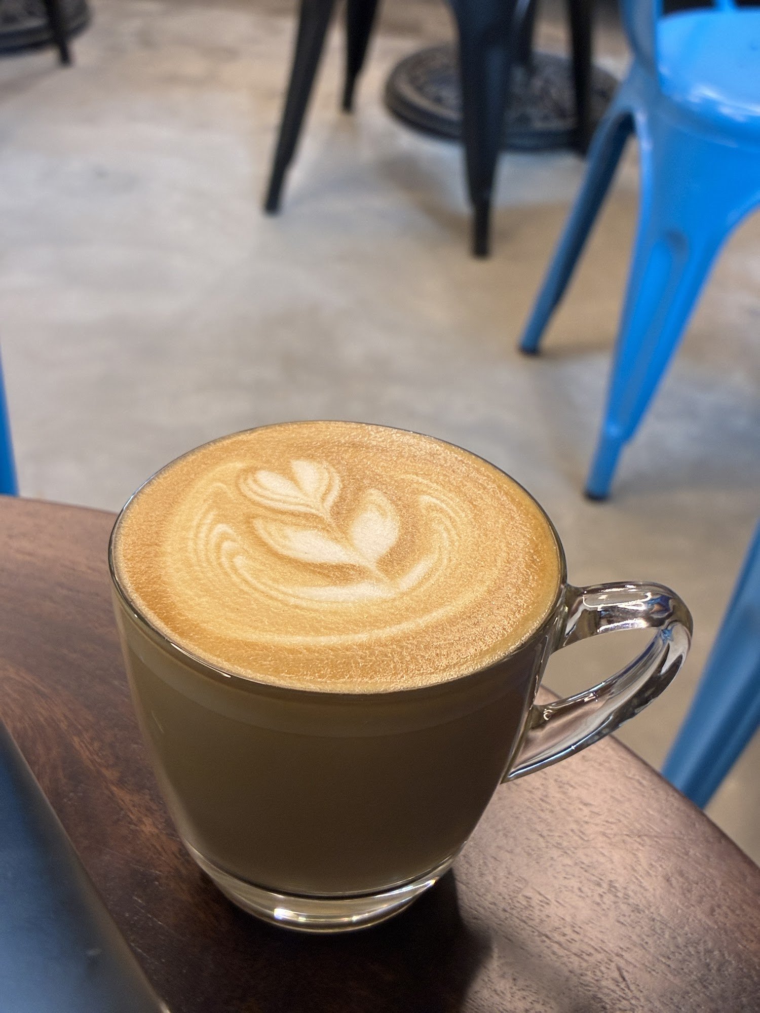 A close-up shot of a latte in a metallic cup on a wooden table at an indoor venue with blue chairs and tables visible in the background.