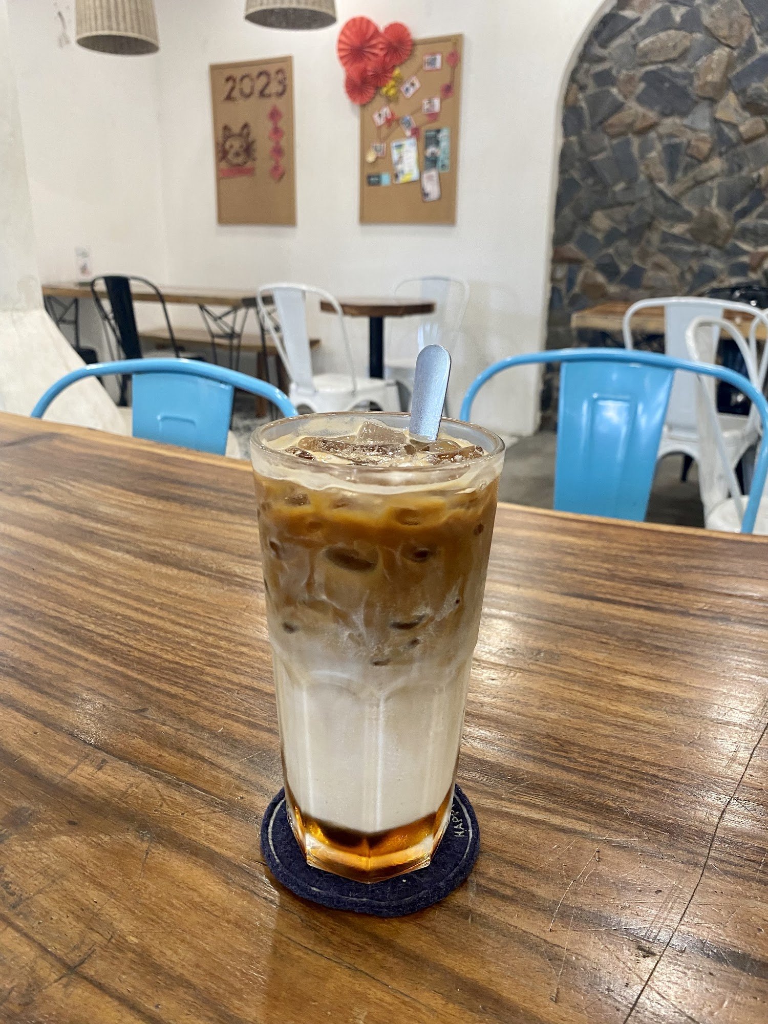 A glass of cold coffee beverage sits on a wooden table in an indoor setting that appears to be a cafe or restaurant with modern decor, including blue 
