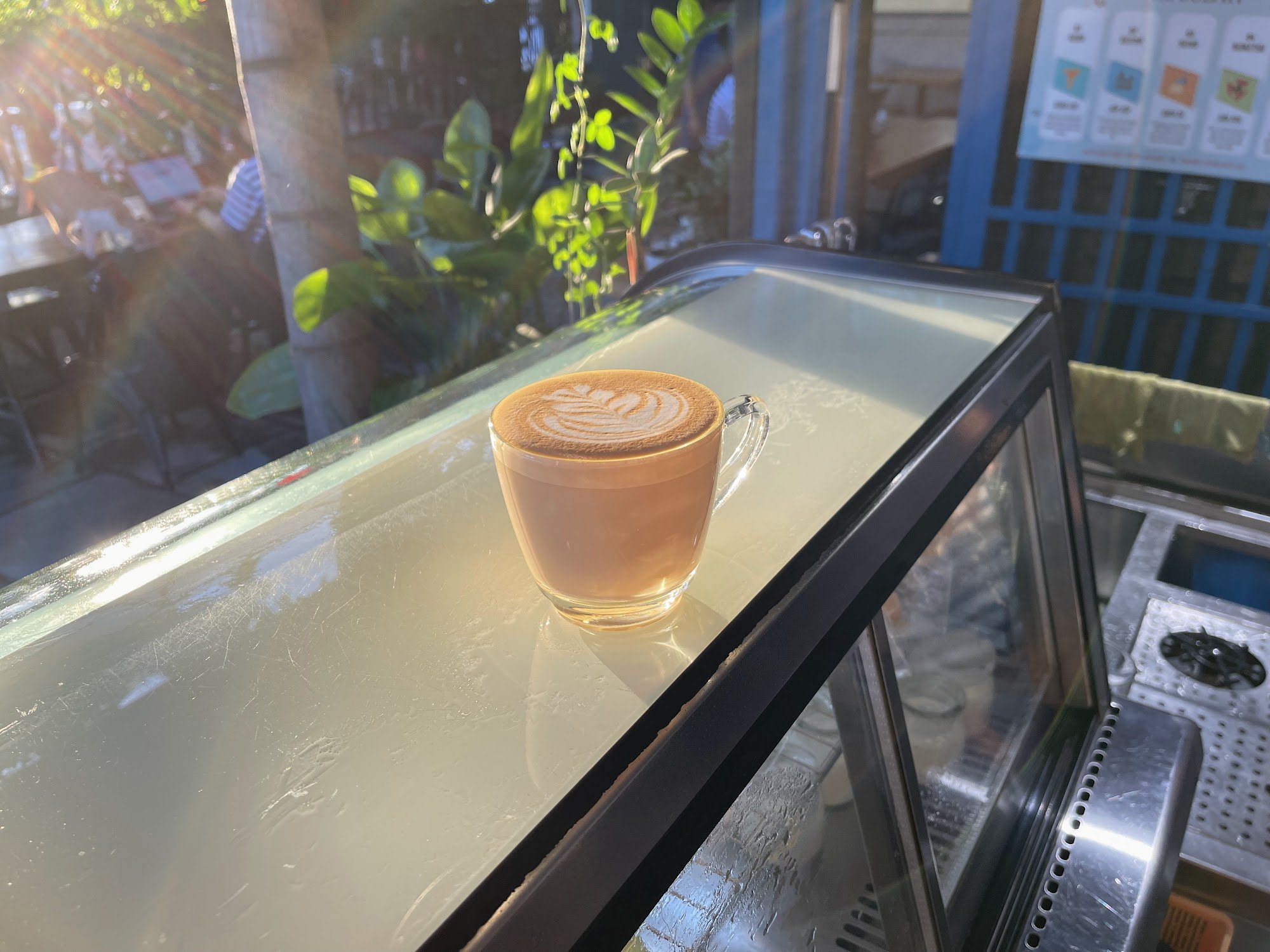 A close-up shot of a latte art coffee on top of a glass mug placed inside an open display case in what appears to be a café setting, with sunlight str