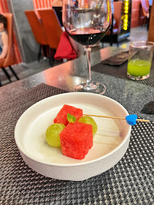 A close-up of a plate containing watermelon and green grapes on a table in an indoor setting with red wine glasses visible in the background.