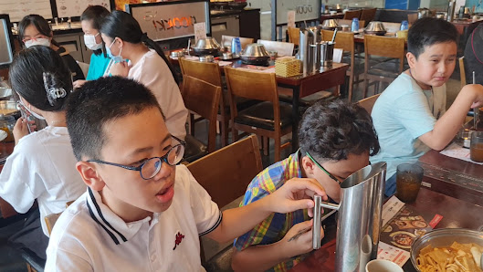 A bustling restaurant interior in Thao Dien, Saigon, with multiple patrons seated at wooden tables. People are engaged in eating, some using utensils,