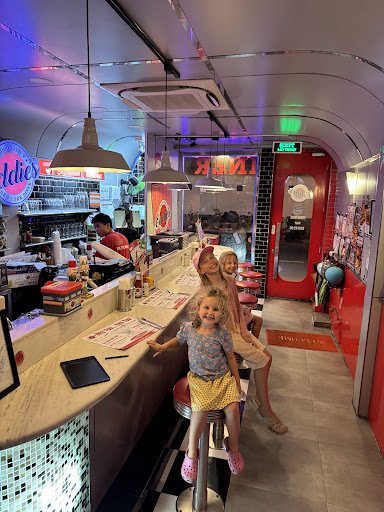 The image shows an interior view of a retro-themed diner named 'Oldies'. The decor includes neon signs and vintage-style lighting fixtures, creating a