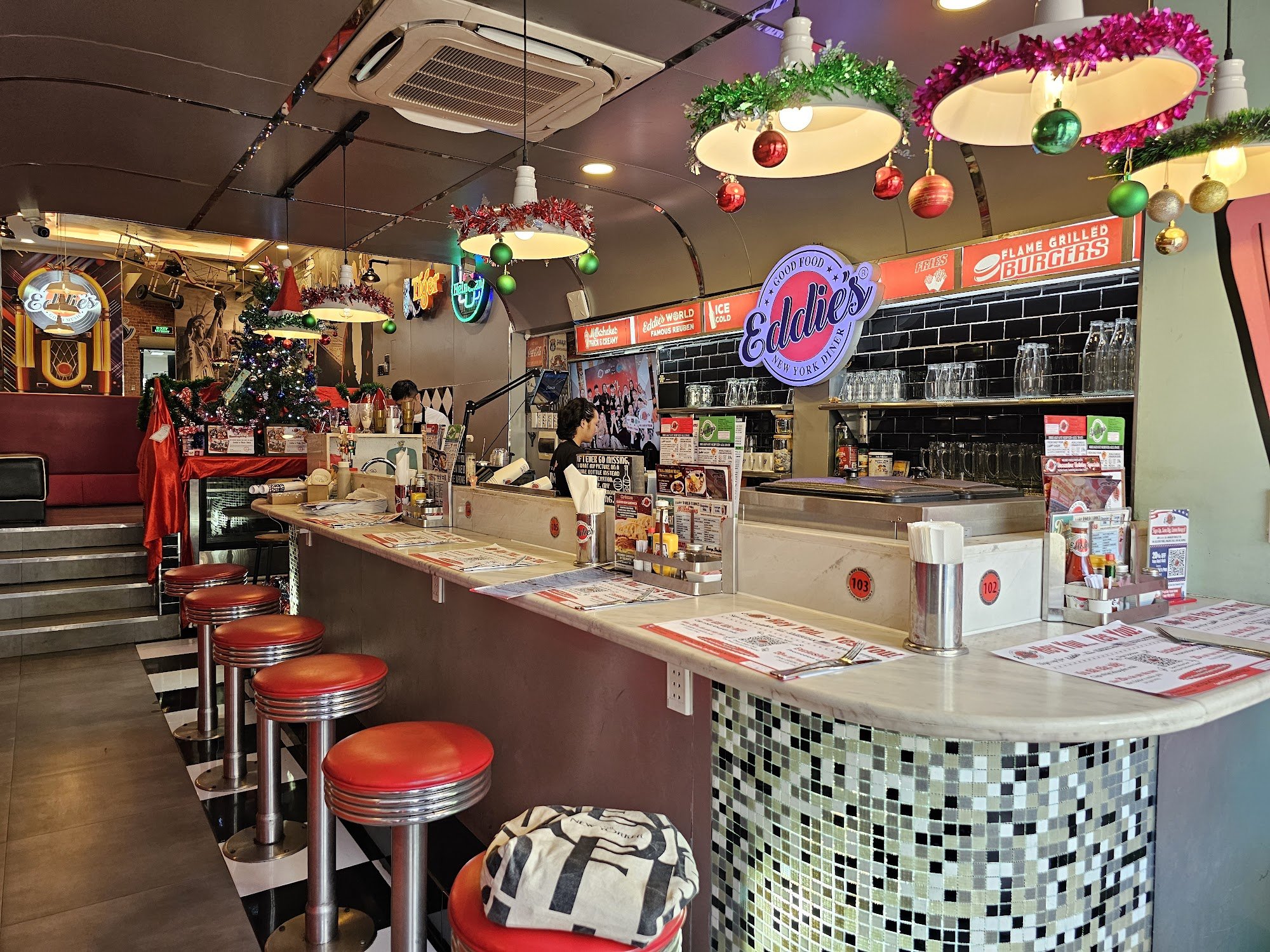 The image depicts the interior of Eddlie's New York Diner in Thao Dien, Saigon. It features a retro-style diner counter with red cushioned bar stools,