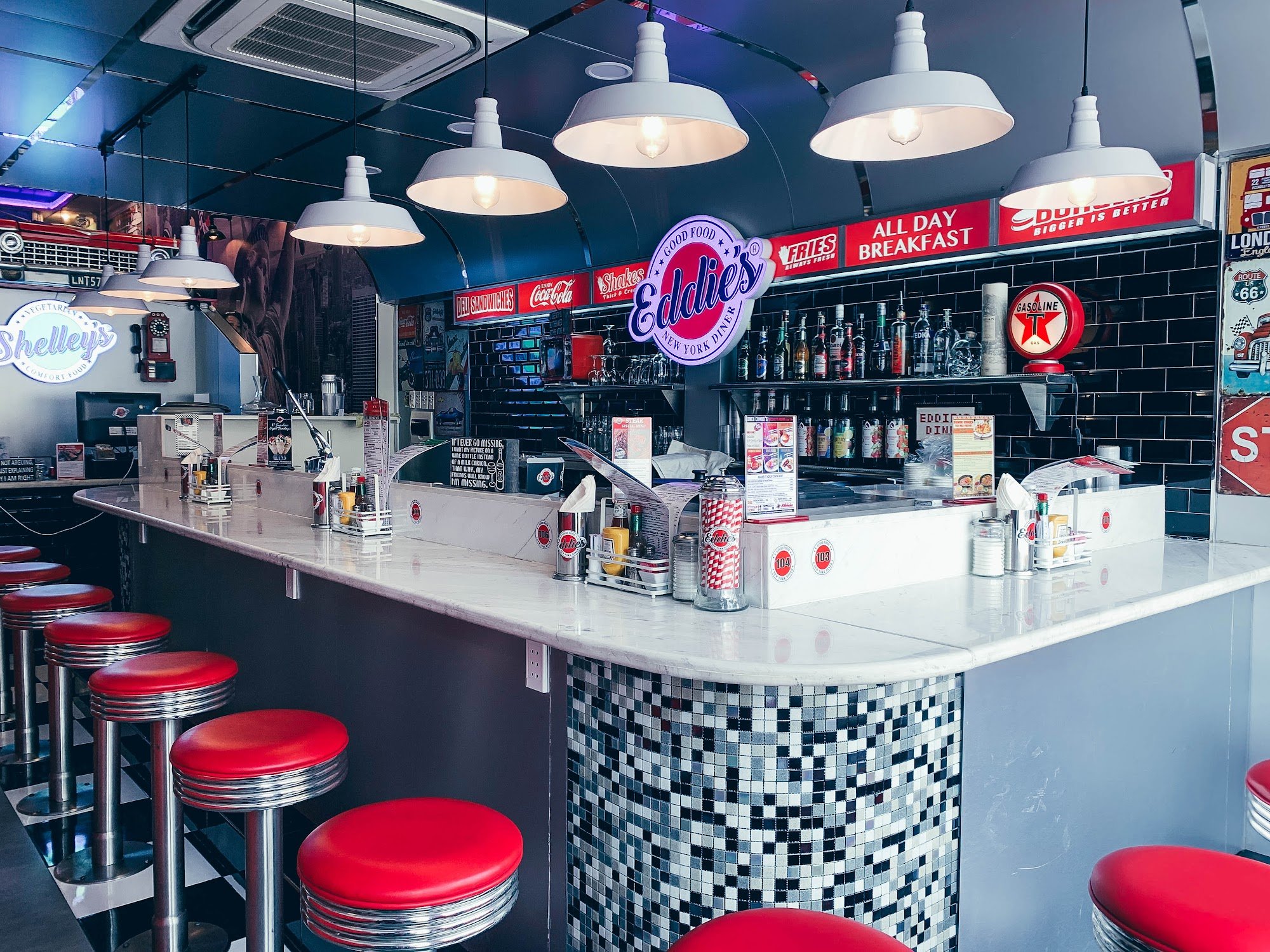 Interior of a retro-style diner named 'Eddie's New York Diner' with a curved counter, red cushioned bar stools, white pendant lights, black tile backs