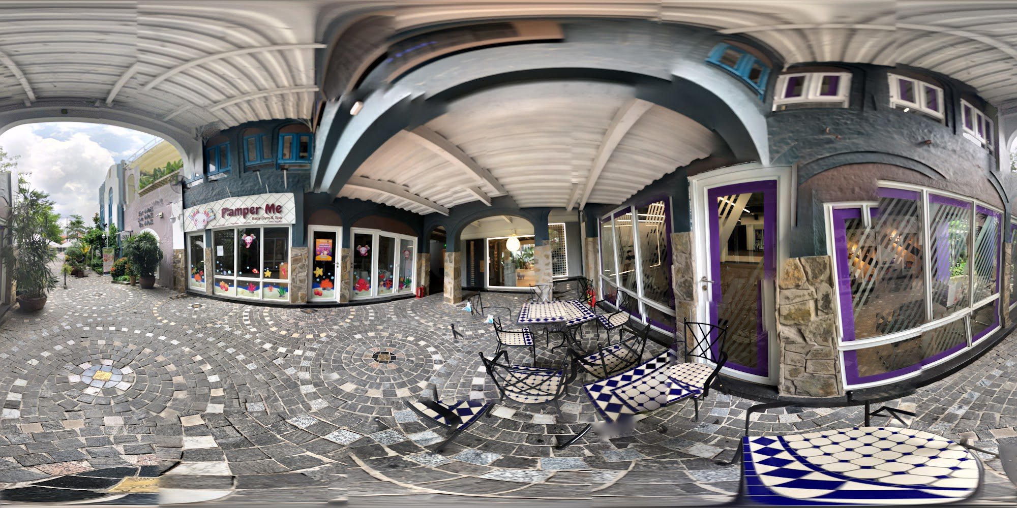 The image shows a panoramic view of an outdoor seating area in front of a restaurant named 'Pamper Me'. The setting includes decorative pavement, blue