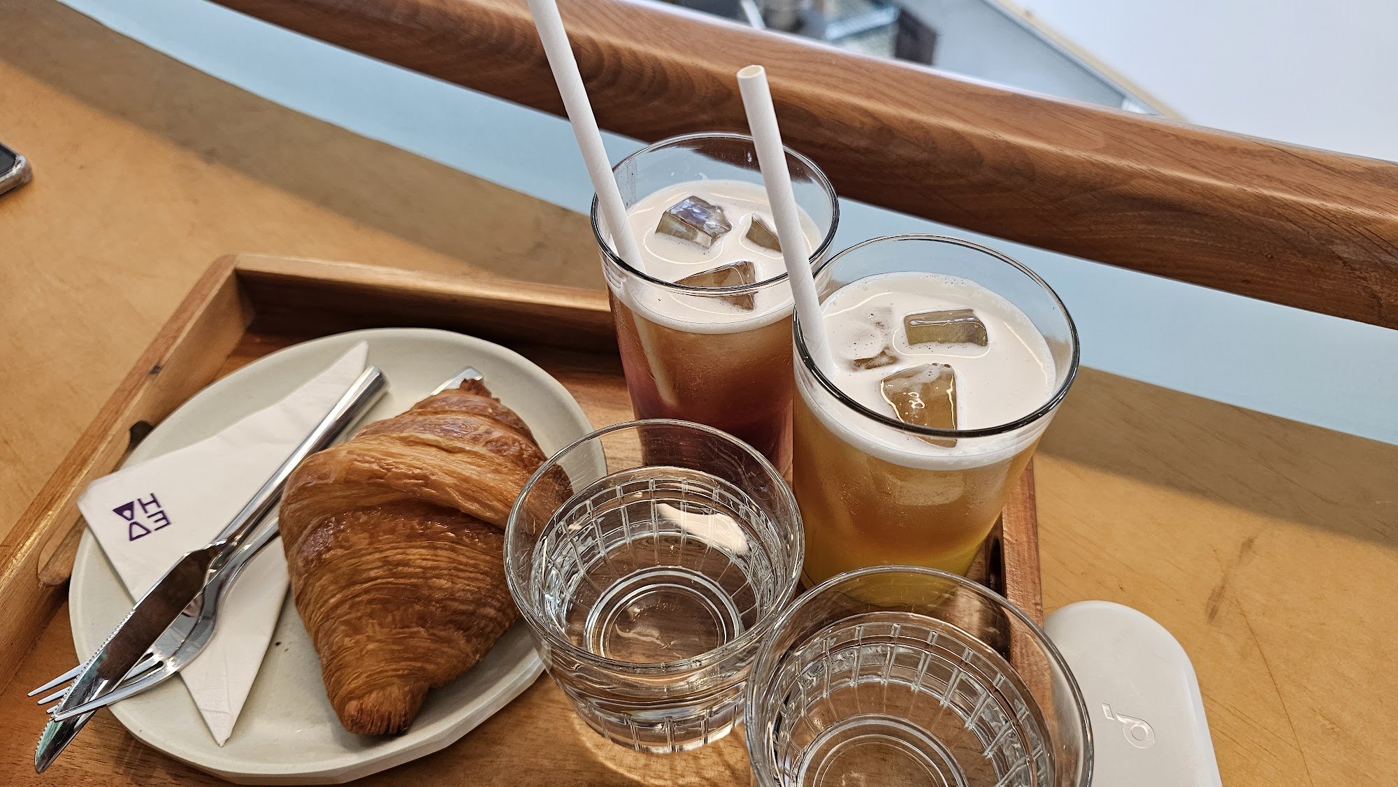 The image shows a wooden tray on which there are two glasses of frothy beverages with ice cubes and straws, another glass that appears to be empty, a 
