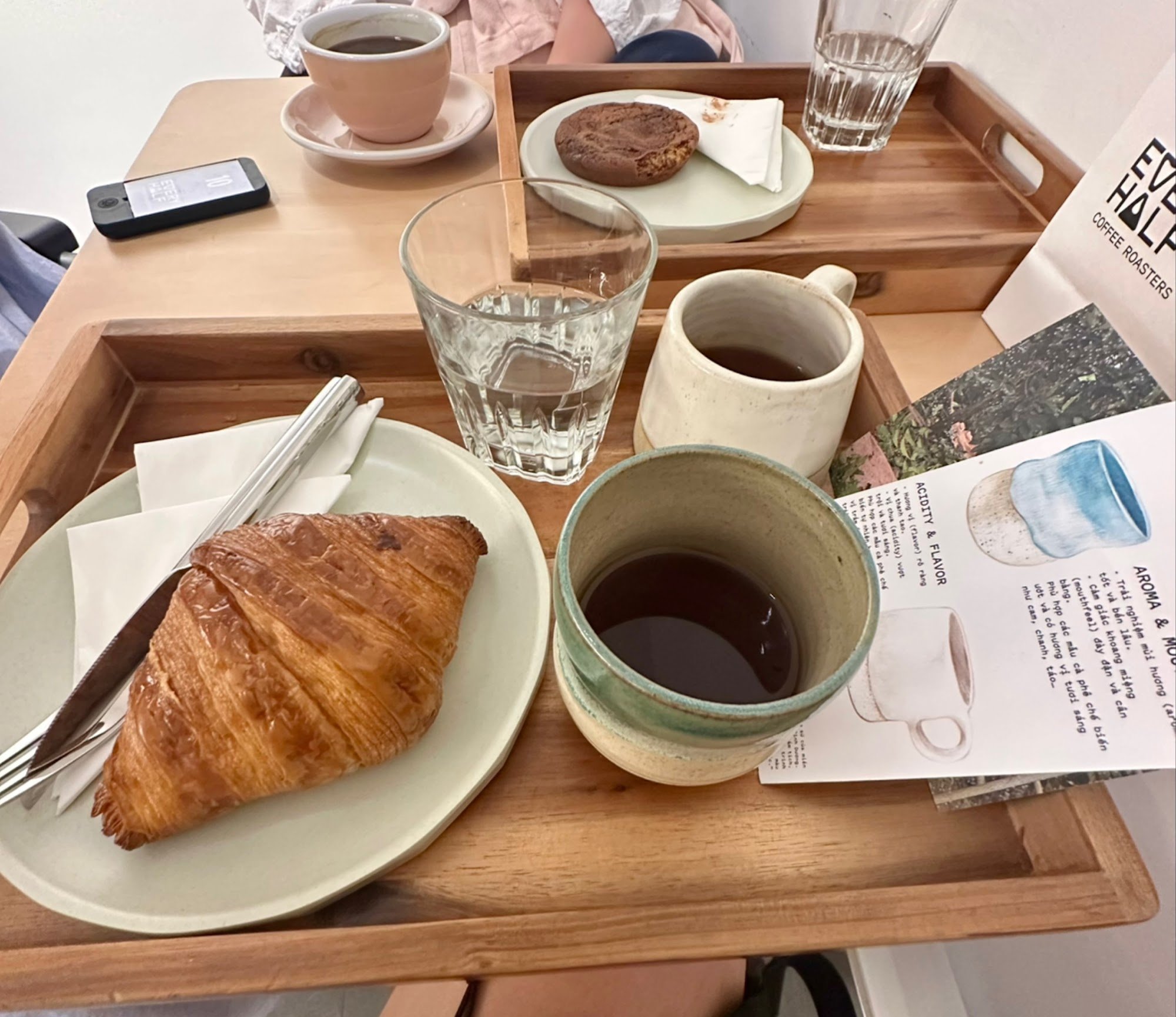 A breakfast setting on a wooden tray includes a croissant, coffee in various cups and glasses, water, cookies, and reading material.