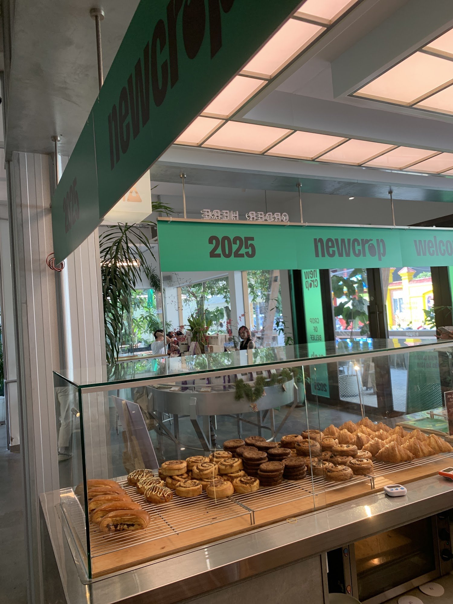 The image depicts the interior of a café named 'newcrop' in Thao Dien, Saigon. A glass display case showcases various pastries including croissants, c