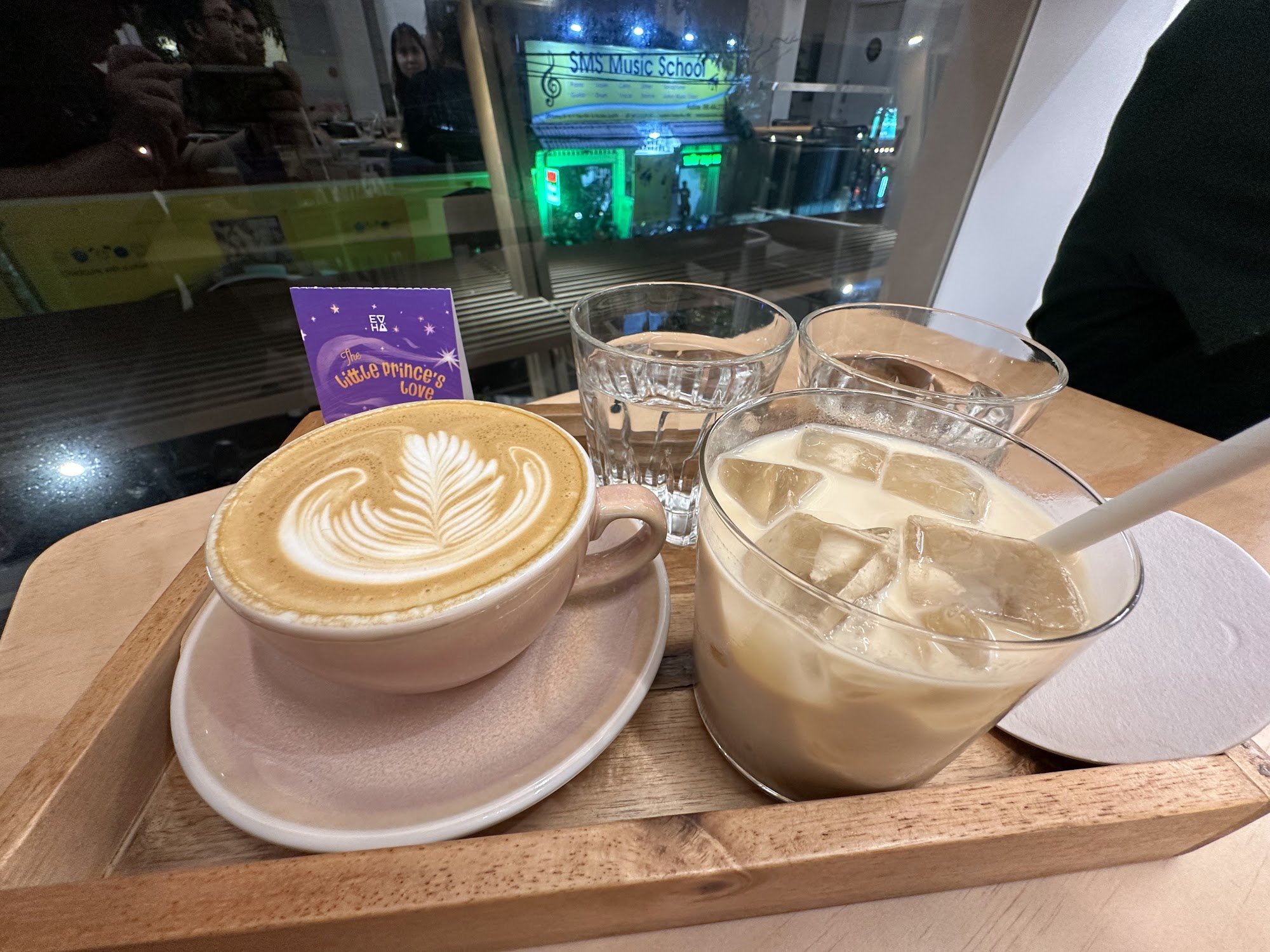 A wooden tray holds a variety of beverages including two cappuccinos and an iced coffee drink on top of it, with a glass of water beside them. In the 