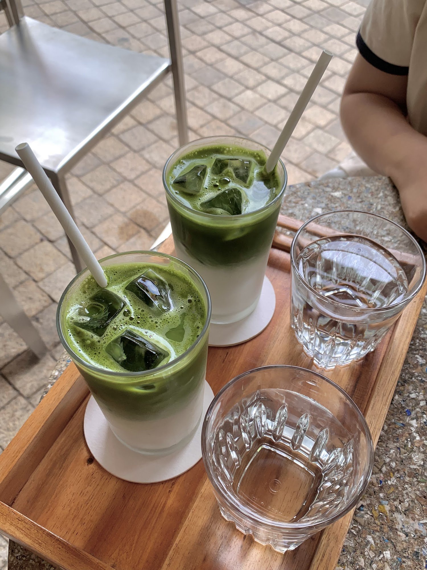 Two green matcha drinks topped with ice and served in clear glasses on a wooden tray alongside two empty drinking glasses.