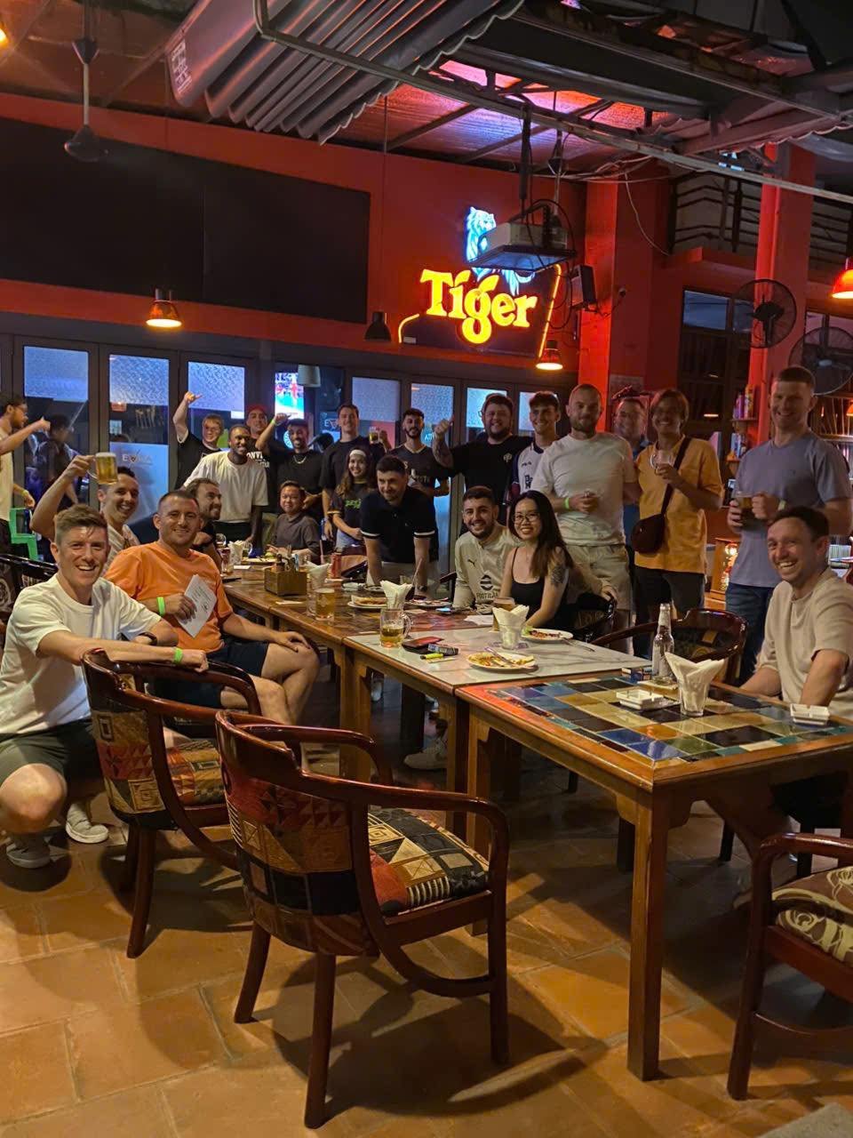 A group of people is gathered in a lively bar setting at Thao Dien, Saigon. They are seated around tables and standing near the counter with drinks in