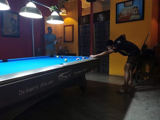 A person is playing pool in a dimly lit room with warm tones and ambient lighting from overhead lamps. The billiards table has the brand 'Atlas' on it